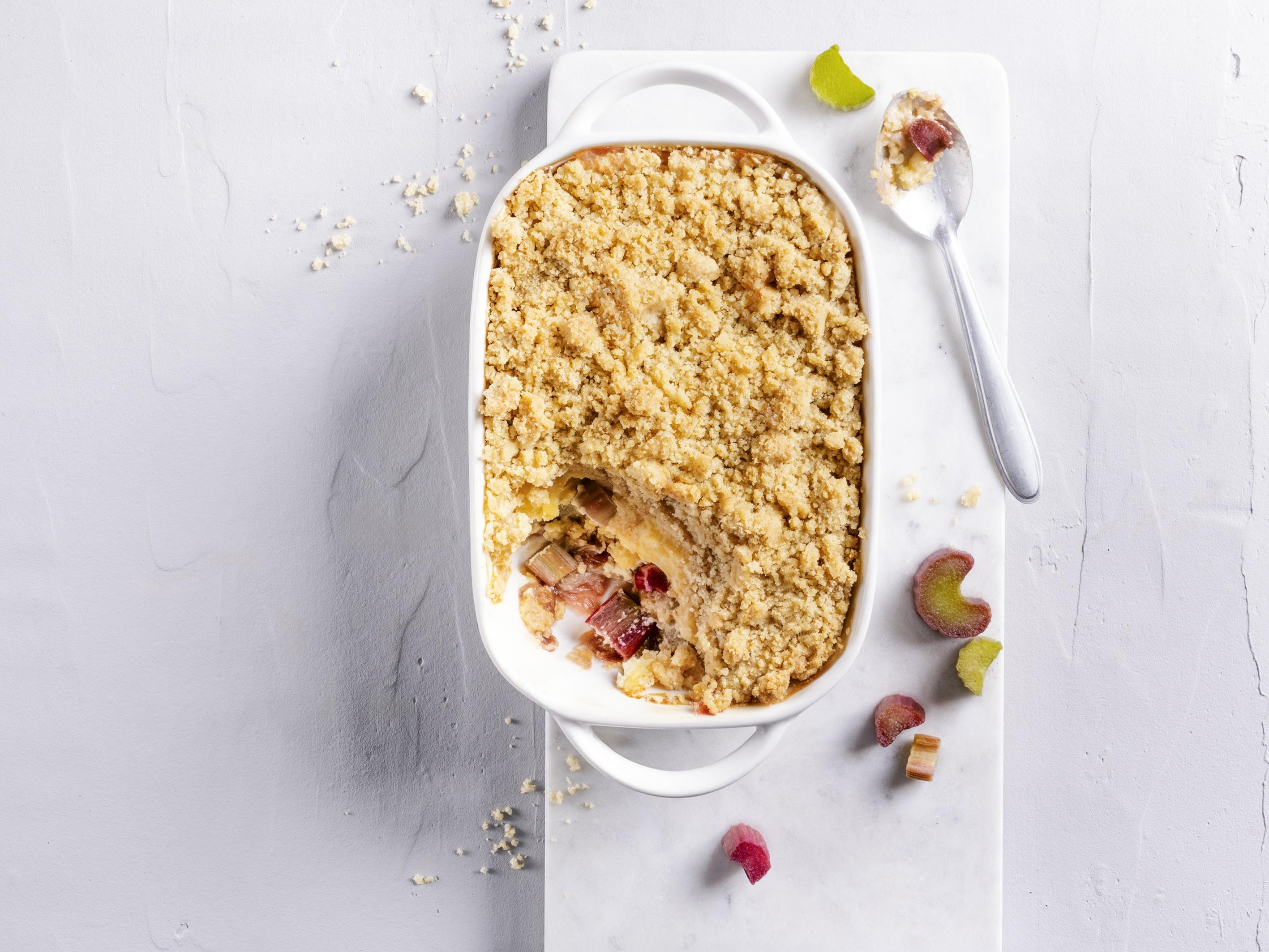 Ein echtes Wohlfühldessert: Rhabarber-Crumble mit Vanille-Créme Eine längliche Auflaufform mit Rhabarbercrumble. Goldgelbe Butterstreusel auf Rhabarber. Rhabarberstückchen als Dekoration.