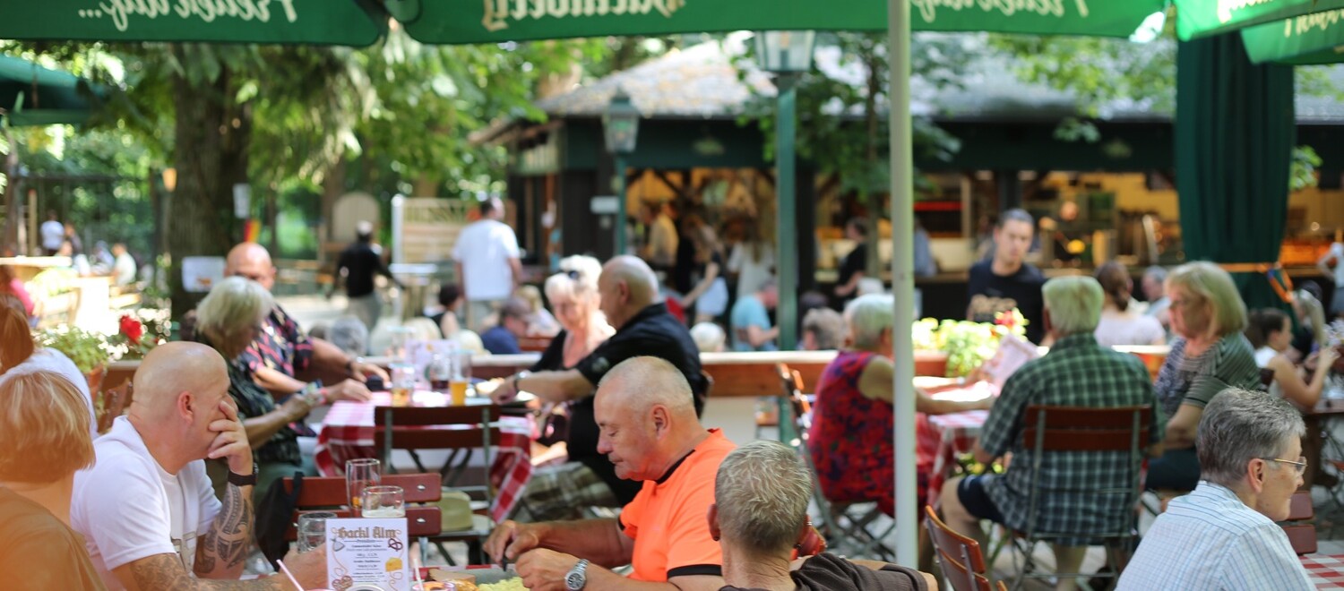 Gäste im Biergarten Eine Gruppe von Menschen sitzt an Tischen unter Sonnenschirmen.