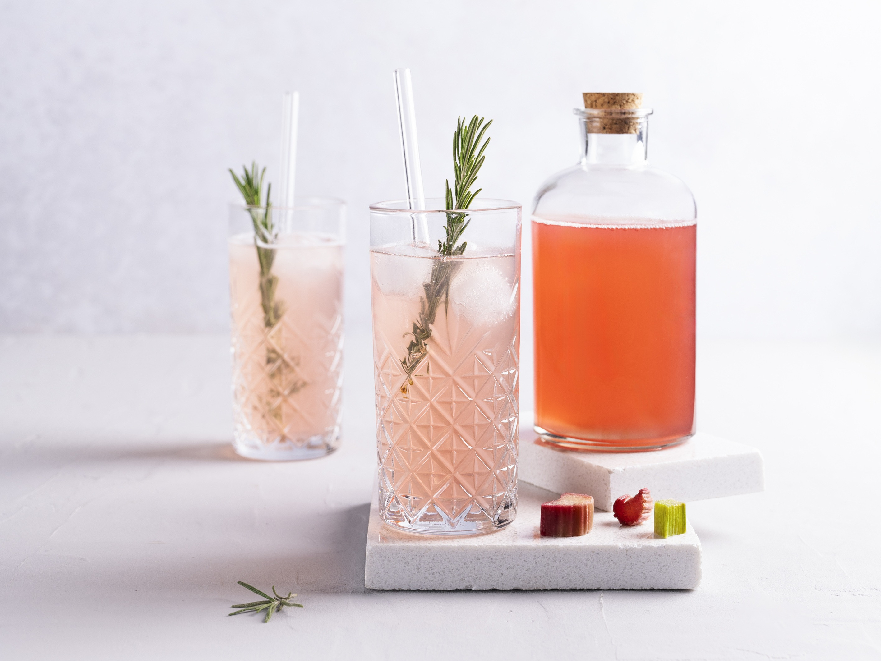 Erfrischende Limonade mit Rhabarber-Sirup Eine Glasflasche mit Rhabarber-Sirup, daneben zwei Kristallgläser mit Rhabarberlimonade, Rosmarinzweige und Rhabarberstückchen als Dekoration