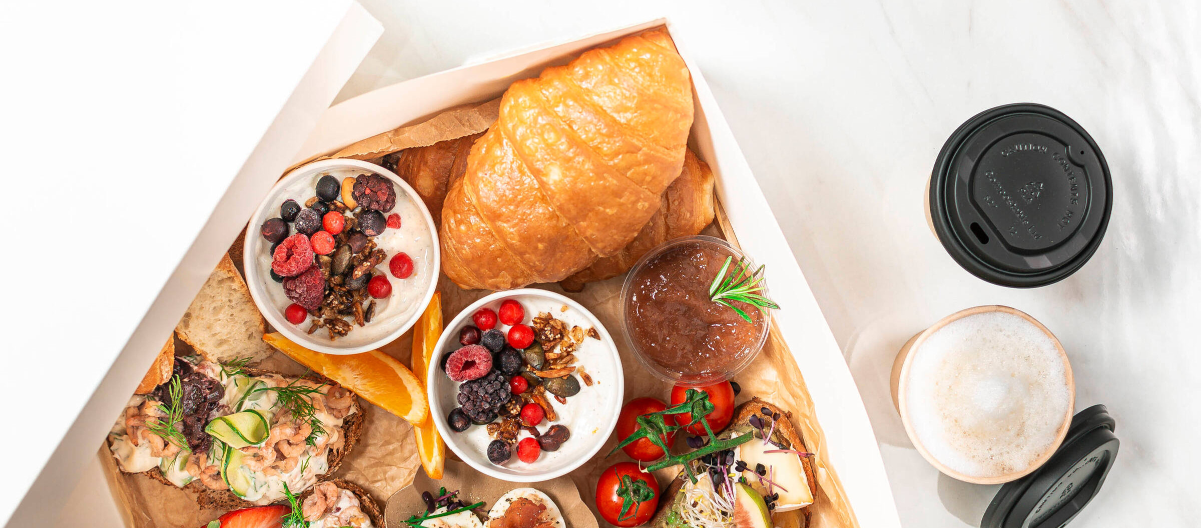 Eine weiße Frühstücksbox mit zweimal Porridge mit beeren, Croissant, Marmelade und einem Coffee to go.