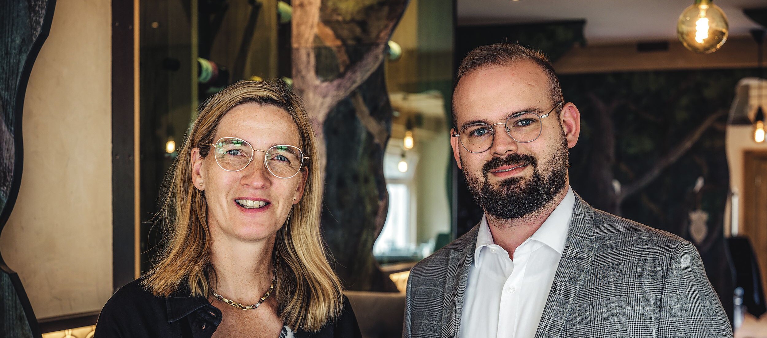 Ein Mann und eine Frau stehen zusammen, beide lächeln, der Mann trägt einen Anzug und eine Brille.