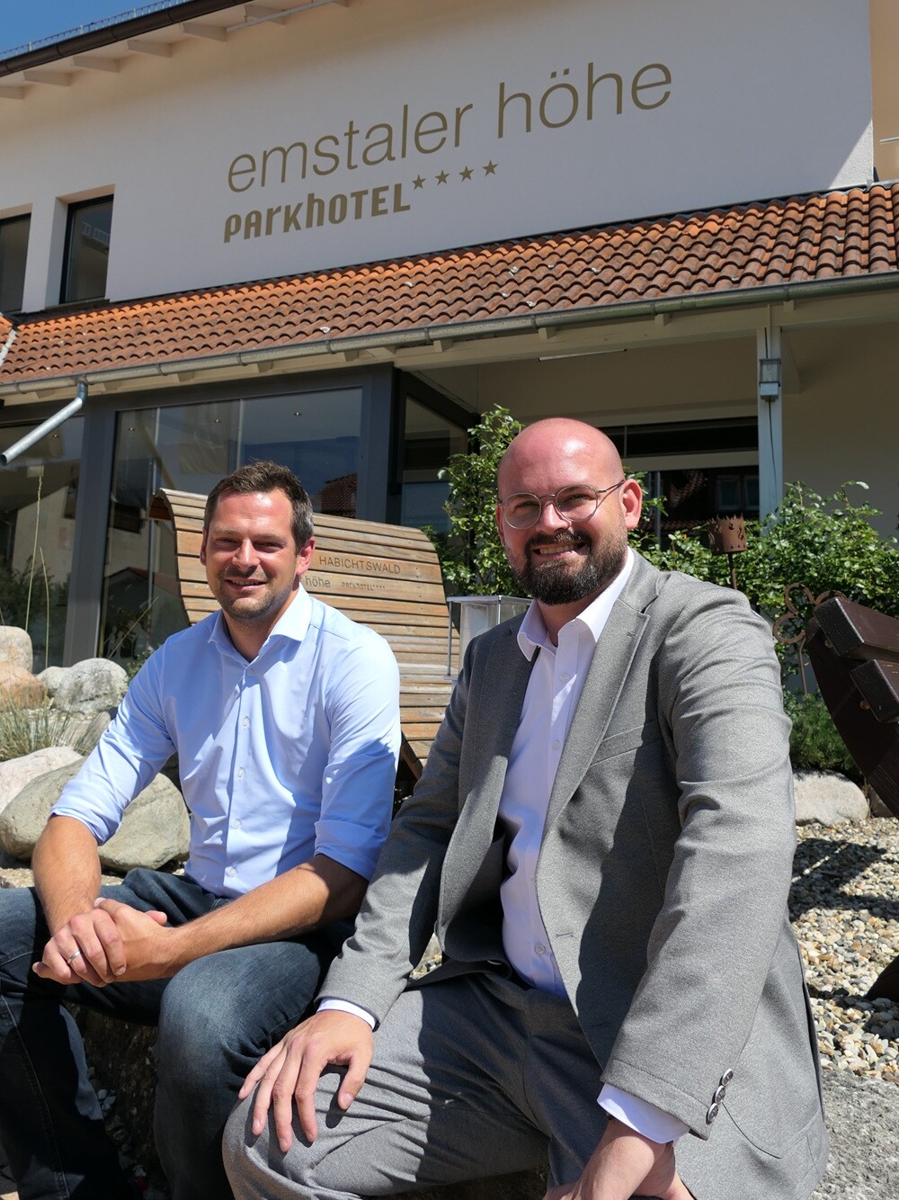 Zwei Männer sitzen lächelnd vor einem Gebäude.