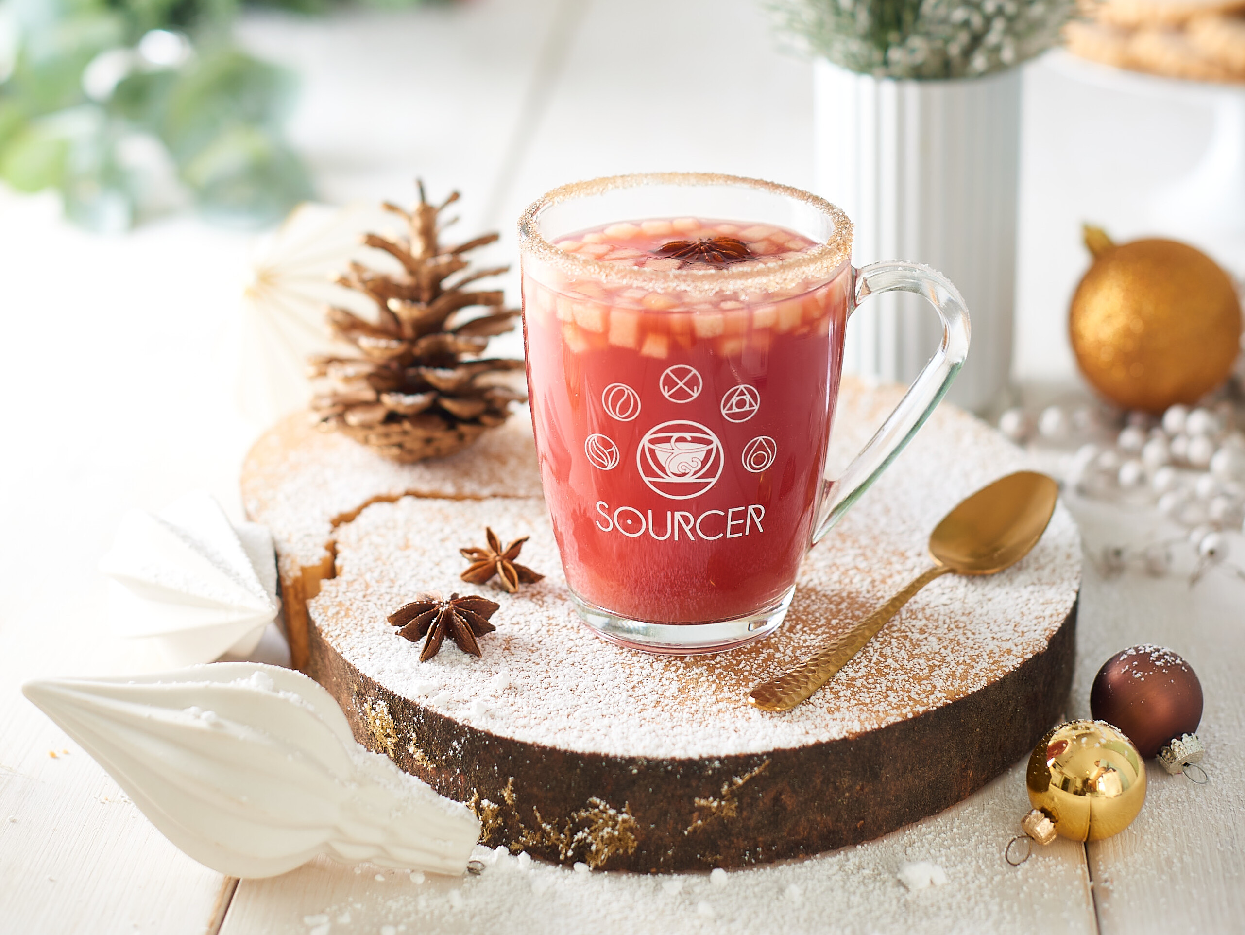 Ein Glas mit roter Flüssigkeit auf einer Holzscheibe, daneben ein Löffel und ein Tannenzapfen.