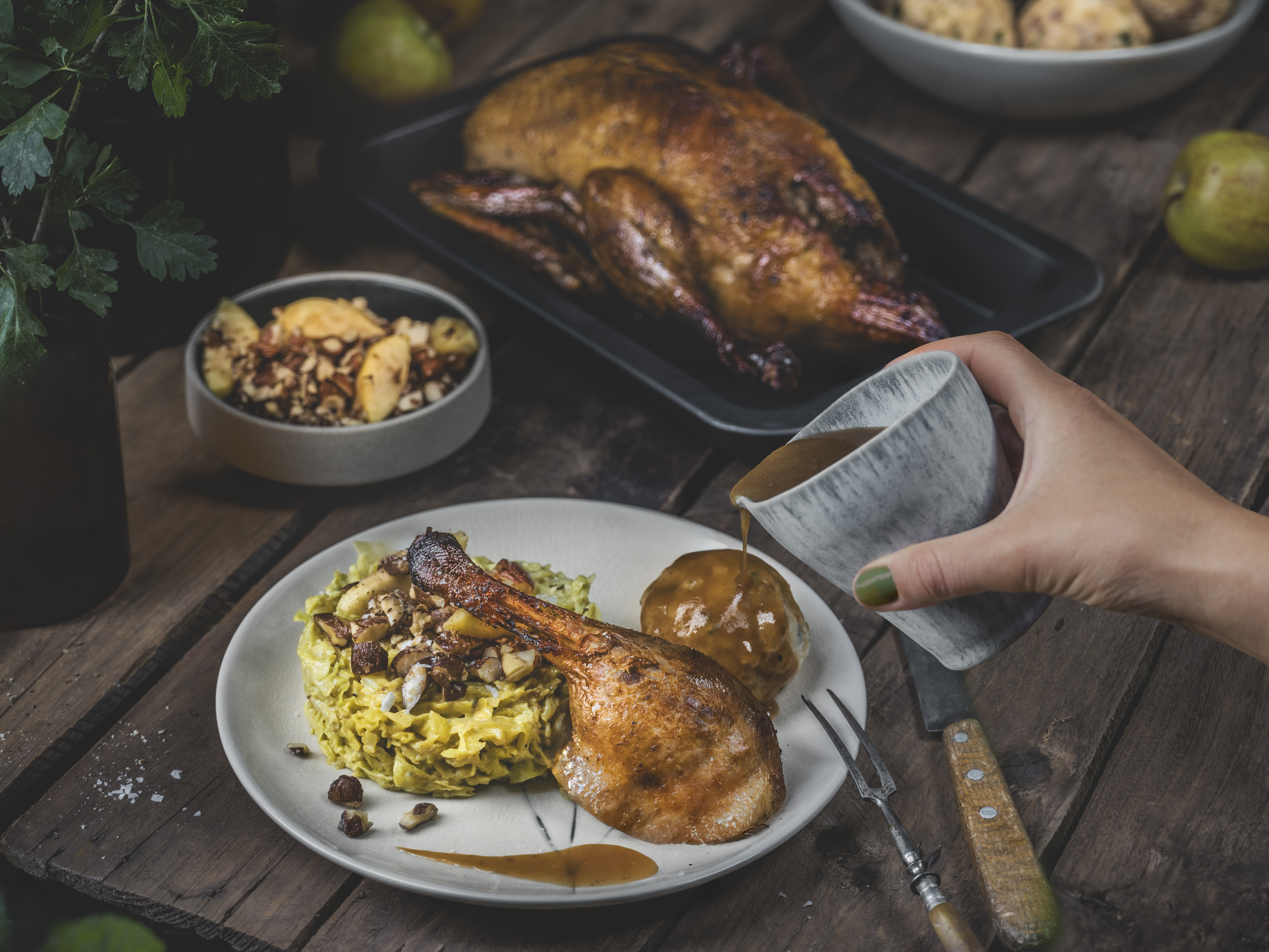 Keule von der Bauernente mit Wirsing und Tiroler Speckknödel. Eine gebackene Entenkeule mit Wirsing-Gemüse und einem Knödel auf einem Teller angerichtet. Eine Hand gießt Soße über den Knödel. Im Hintergrund liegt eine ganze Bauernente.