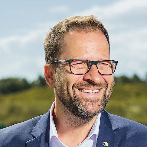 Ein Mann im Anzug lehnt an einem Geländer und lächelt.