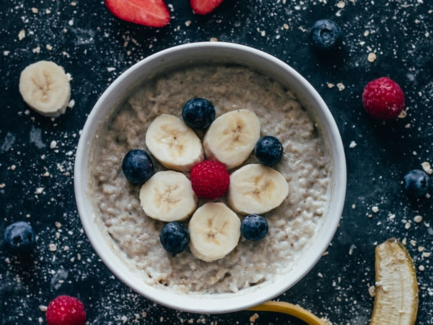 Porridge mit Blaubeeren, Bananenscheiben und einer Himbeere in einer weißen Schale auf einer dunklen Schieferplatte mit Puderzuckersprenkeln.