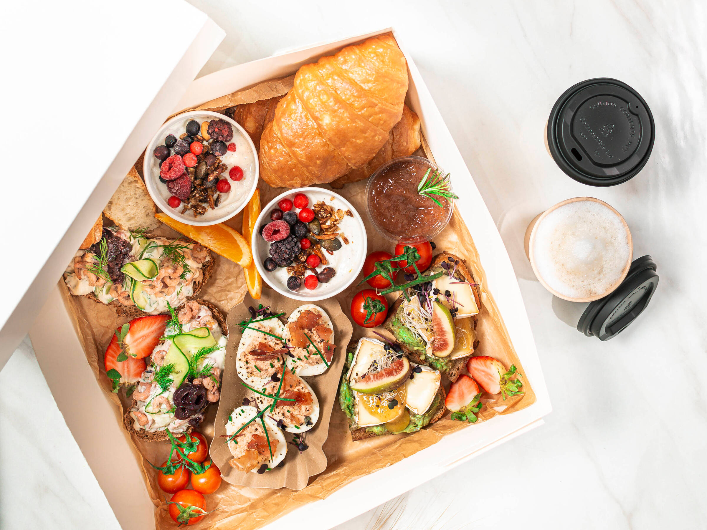 Eine Frühstücksbox mit Croissants, Porridge, belegten brötchen, Eiern und Obst und Gemüse. Außerdem Kaffee in zwei Togo-Bechern an der Seite.